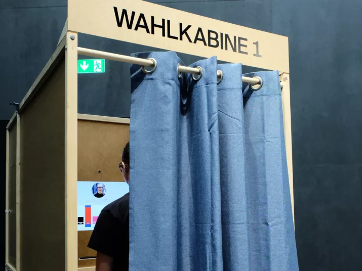 Ein Mensch steht in einer Wahlkabine, der Vorhang ist halb zugezogen.